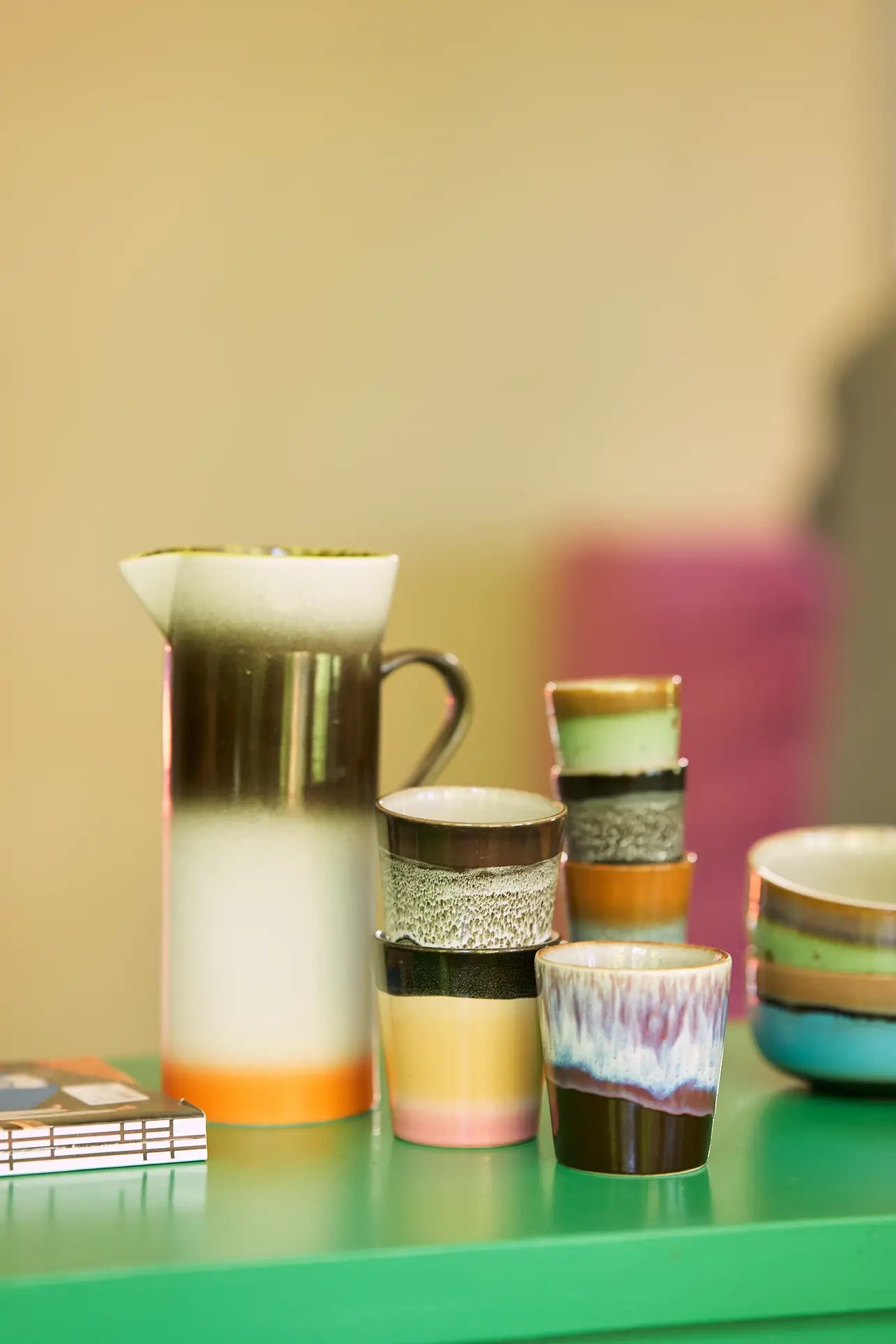 The HKLIVING 70s Ceramics Coffee Mugs, Set of 6, with handcrafted glazing, are displayed on a green tabletop with a blurred background and a small stack of books in the corner.