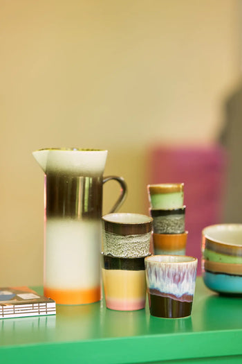 The HKLIVING 70s Ceramics Coffee Mugs, Set of 6, with handcrafted glazing, are displayed on a green tabletop with a blurred background and a small stack of books in the corner.