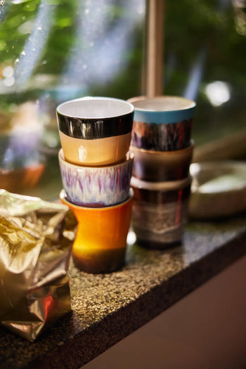 A set of HKLIVING 70s Ceramics Coffee Mugs (set of 6) with handcrafted glazing sits on a kitchen windowsill, sunlight streaming in and greenery visible outside, next to a crumpled gold foil package.