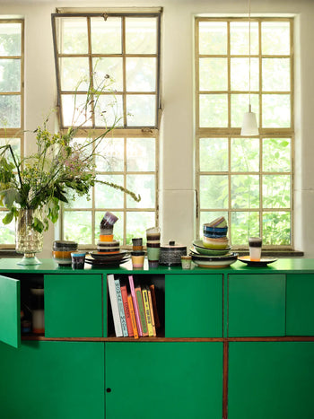 A bright kitchen with large windows, green cabinets, stacked dishes, a vase of wildflowers, colorful books on open shelves, and HKLIVING 70s Ceramics Coffee Mugs set of 6. Sunlight streams in, making the space feel fresh and inviting.
