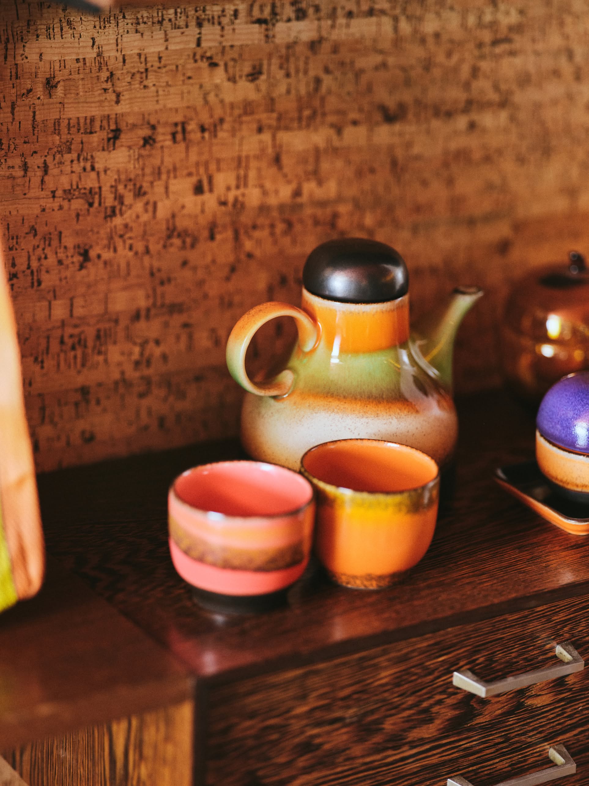 The HKLIVING 70s Ceramics: Coffee Pot - Morning sits next to matching cups on a wood surface, its orange, green, and beige glaze creating a bold retro statement against a warm textured background.