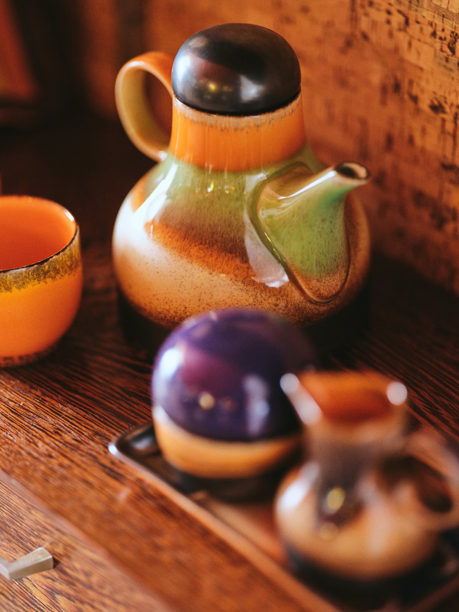 The HKLIVING 70s Ceramics Coffee Pot - Morning, with its green, orange, and brown hues, sits next to an orange cup and matching containers on wood, creating a warm retro scene reminiscent of vintage ceramics.