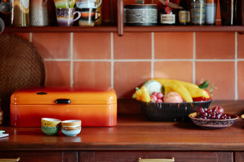 A cozy kitchen countertop with a set of 2 HKliving 70s Ceramics Curry Bowls, an orange bread box, and fresh fruit basket. Above, shelves display mugs and jars, while a warm-toned tile backsplash completes the vintage look.