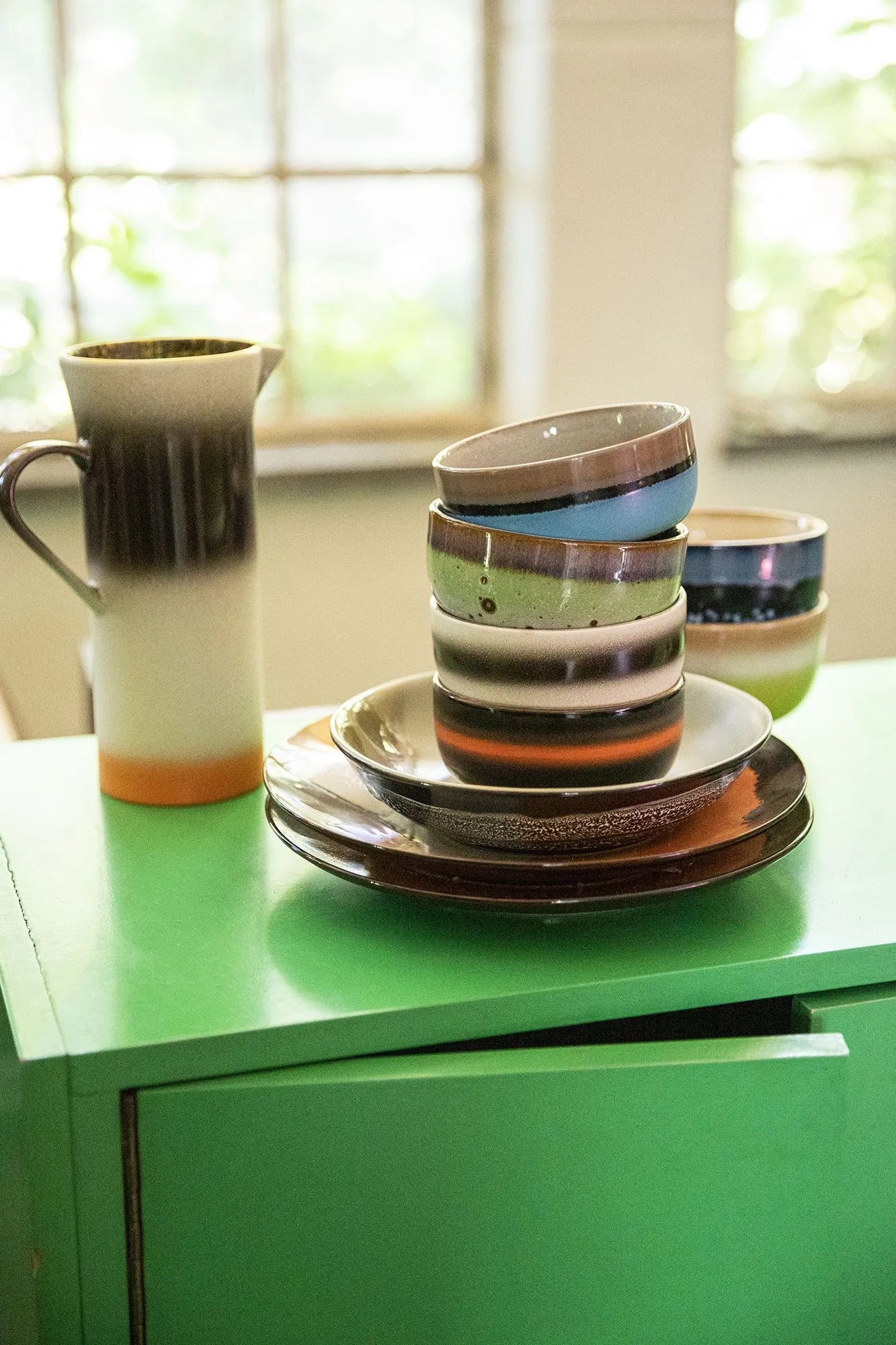 Four HKLIVING 70s Ceramics dessert bowls, featuring earthy multicolored glazes and a retro design, are stacked on a bright green cabinet by a window with blurred greenery outside.
