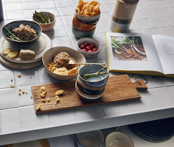 On a tiled kitchen table, HKLIVING 70s Ceramics: Dessert Bowls (Set of 4) are filled with noodles, cherry tomatoes, cornflakes, ginger, and asparagus beside a wooden board, stacked dishes, and an open cookbook displaying colorful vegetables.
