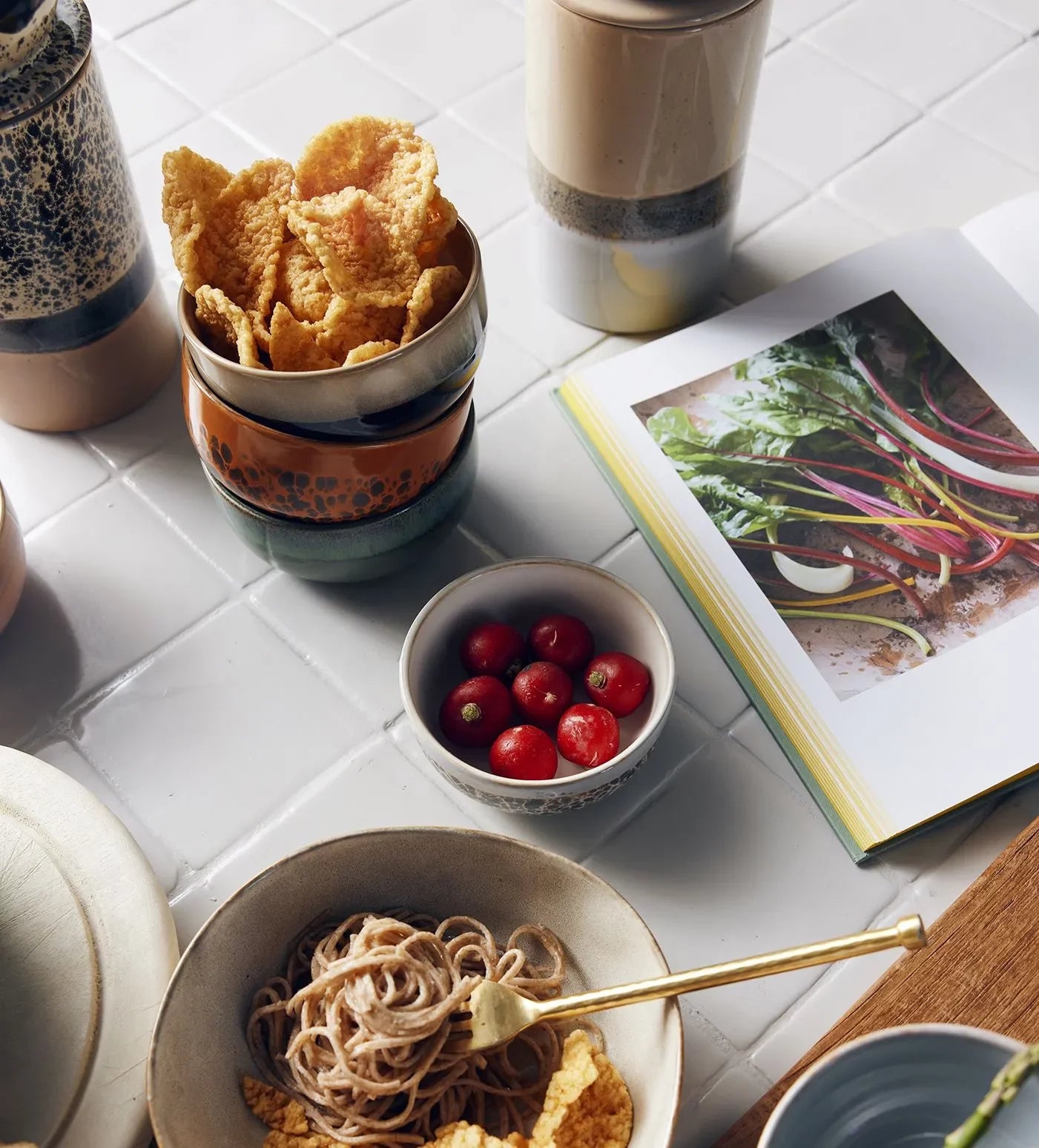 A tabletop styled with charming retro design features HKLIVING 70s Ceramics Dessert Bowls filled with cherry tomatoes and snacks, a bowl of noodles with a gold fork, two mugs, and an open cookbook displaying fresh Swiss chard.