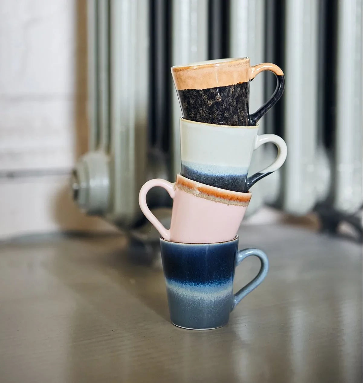 The HKLIVING 70s Ceramics Espresso Mugs, Set of 4, are stacked in a leaning tower on a smooth grey surface, with a radiator visible in the background.