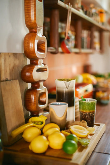 A wooden kitchen countertop holds lemons, a lime, and a banana on a chopping board beside ceramic cups and the HKLIVING 70s Ceramics: Jug - Bomb. In the background, shelves display jars, utensils, and vintage-inspired ceramics by HKLIVING.