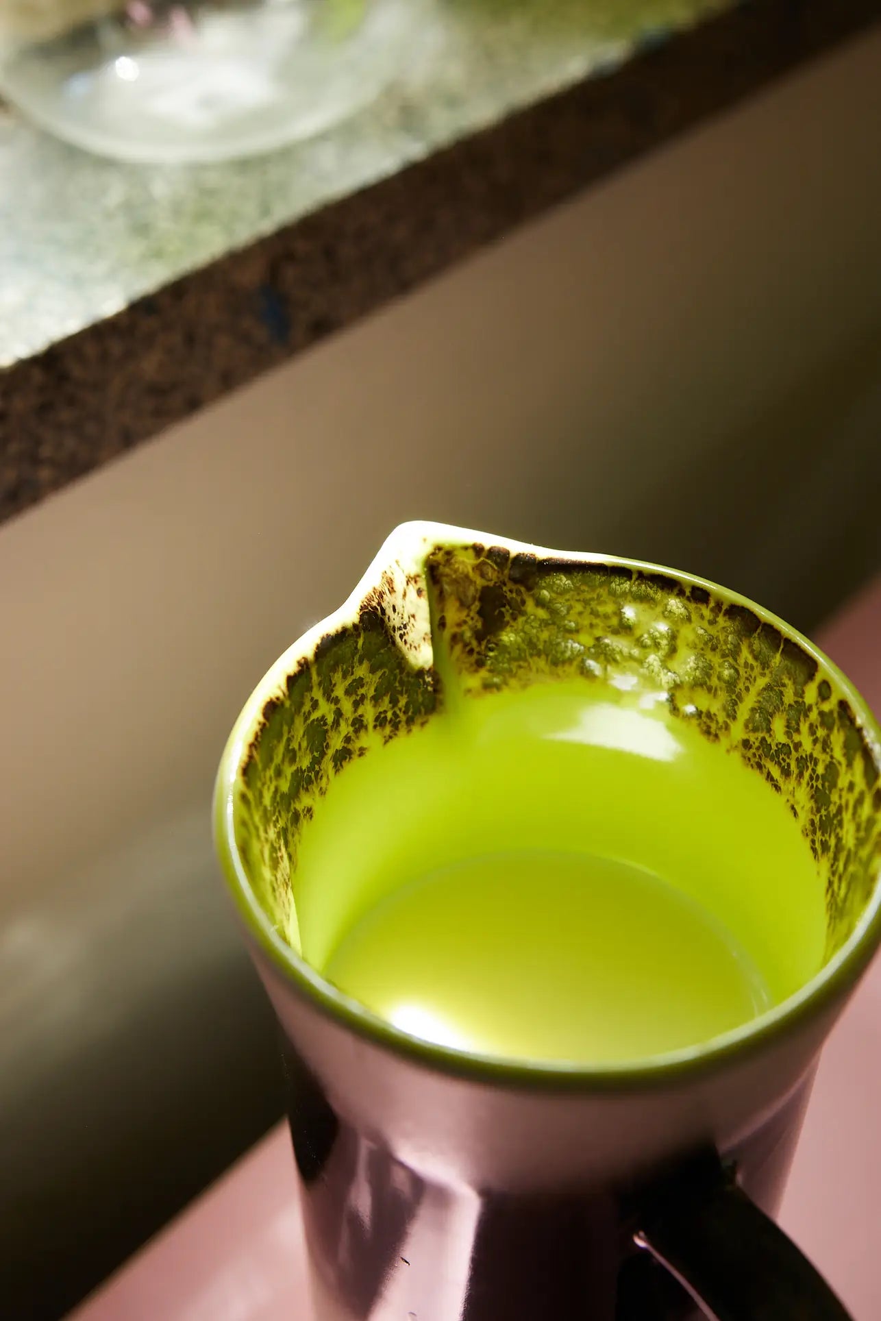 The HKLIVING 70s Ceramics: Jug - Bomb, with a green-tinted retro design and lipped spout, rests on a counter holding yellowish liquid. Stains on its rim hint at past use for frothing or steaming milk.
