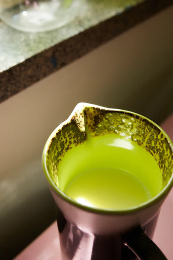 The HKLIVING 70s Ceramics: Jug - Bomb, with a green-tinted retro design and lipped spout, rests on a counter holding yellowish liquid. Stains on its rim hint at past use for frothing or steaming milk.