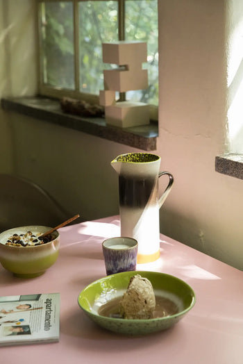 A pink table set with a bowl of granola and yogurt, a modern accent cup, the HKLIVING 70s Ceramics: Jug - Bomb, a green bowl with a scone in sauce, and an open magazine; sunlight streams in through the window.
