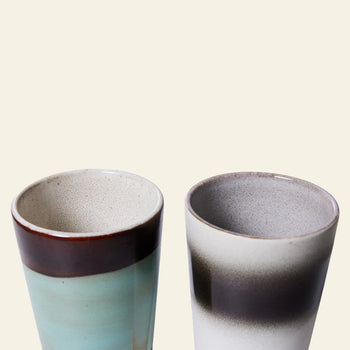 HKLIVING 70s Ceramics: Latte Mugs, Set of 2 by HKLIVING features one turquoise mug with a brown rim and one off-white mug with a dark gray band. Both have matte speckled interiors, echoing retro stoneware mugs from the 1970s.