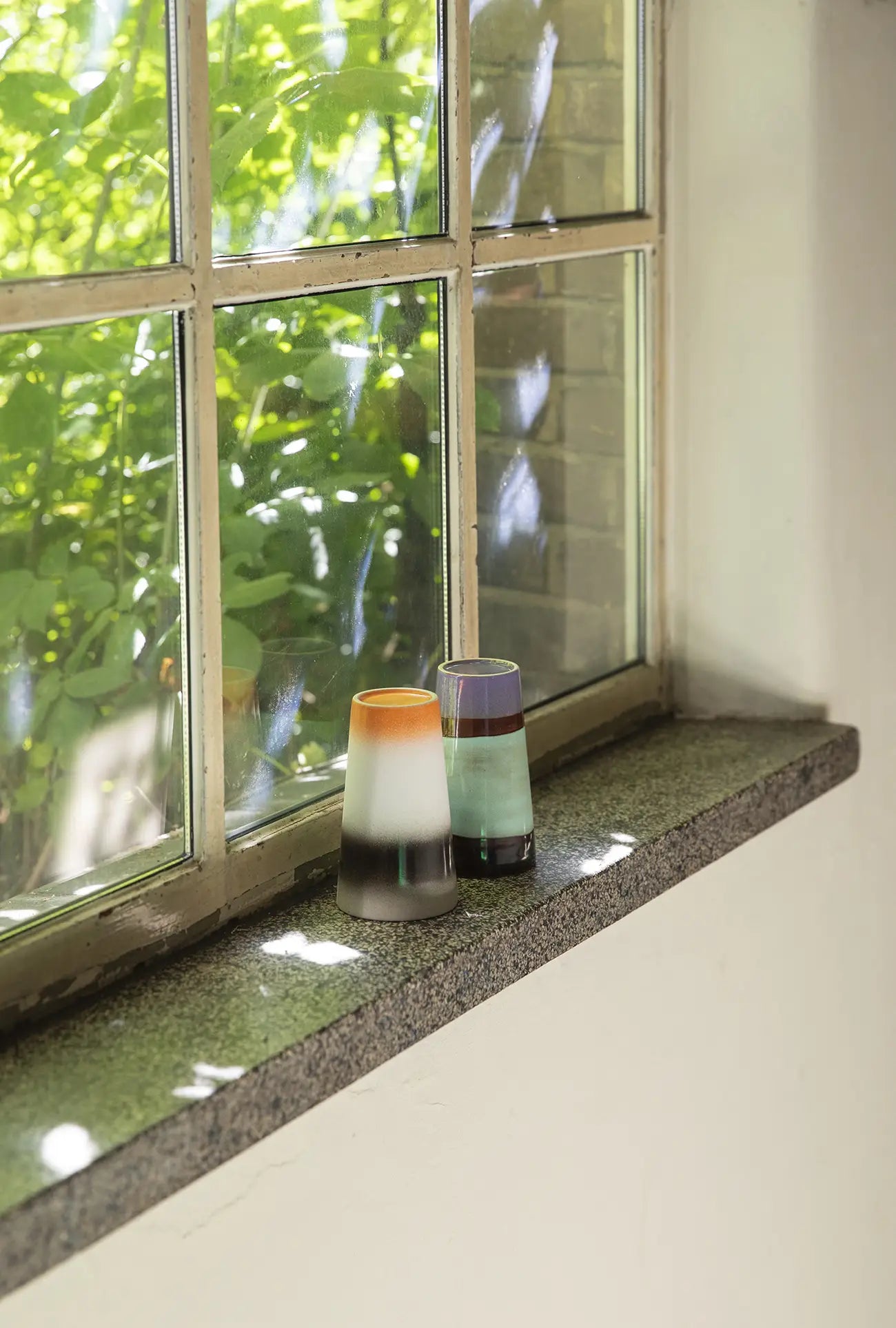 Sunlight streams through two vibrant glass vases on a windowsill, casting shadows. Nearby, HKLIVING 70s Ceramics: Latte Mugs, Set of 2 add retro charm from HKLIVING, with green leaves and branches visible outside.