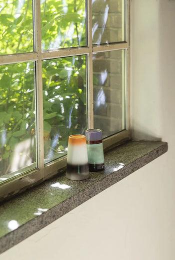 Sunlight streams through two vibrant glass vases on a windowsill, casting shadows. Nearby, HKLIVING 70s Ceramics: Latte Mugs, Set of 2 add retro charm from HKLIVING, with green leaves and branches visible outside.
