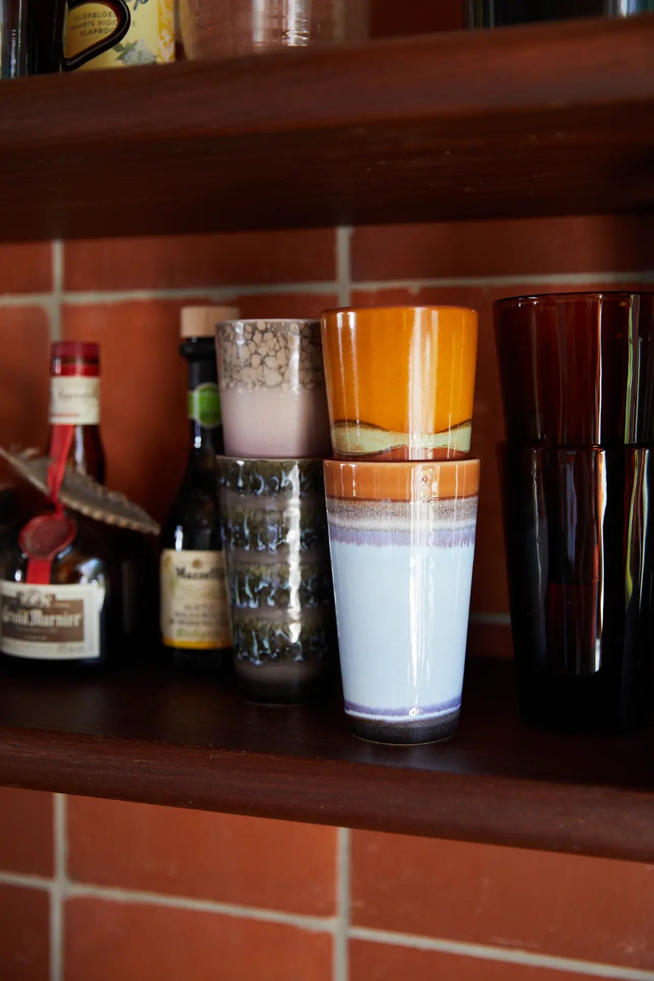 A wooden shelf displays assorted glasses and bottles, including a set of two HKLIVING 70s Ceramics Latte Mugs with orange and blue patterns, dark tumblers, and small bottles, all set against a red tiled wall.