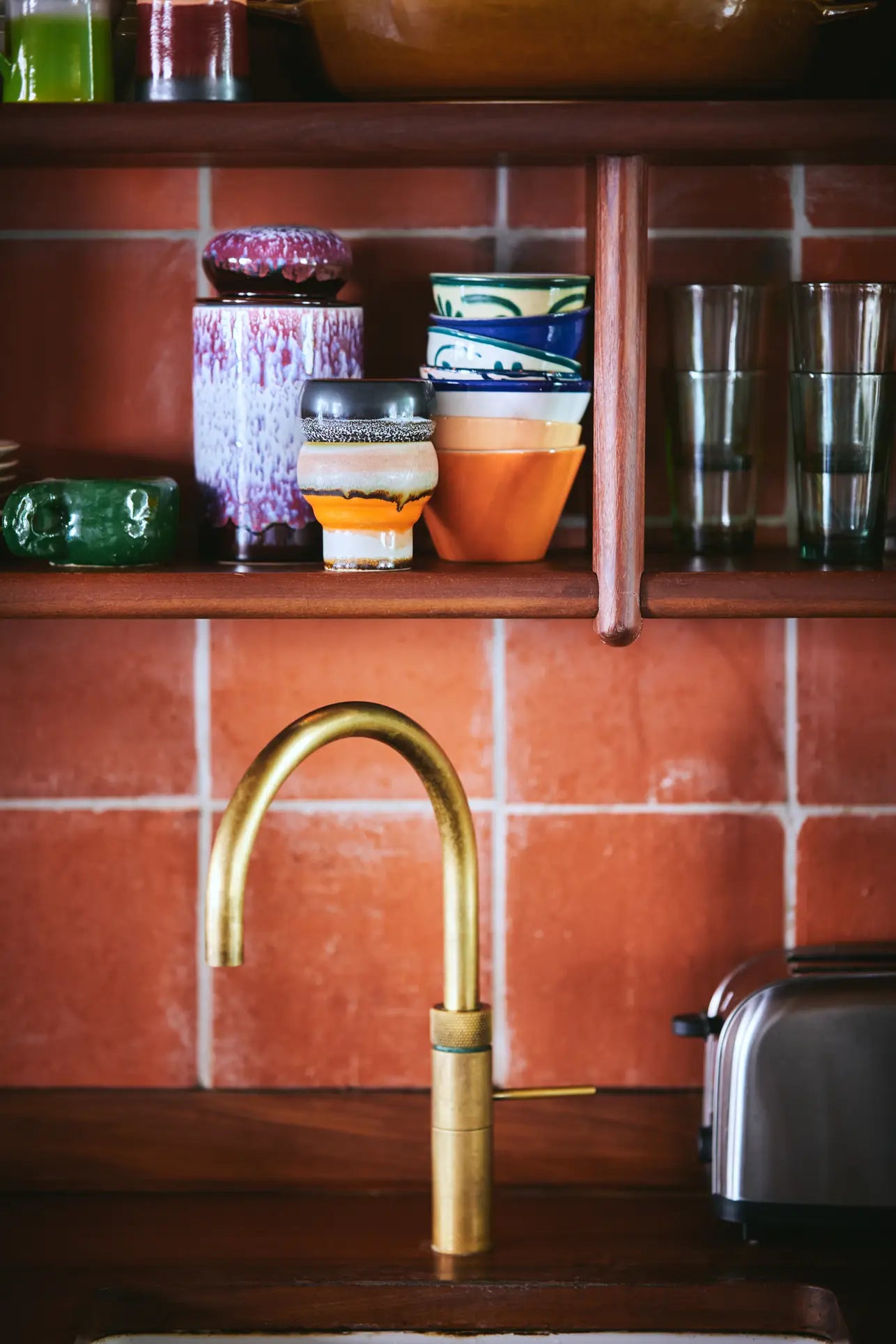 A brass kitchen faucet sits beneath wooden shelves displaying colorful bowls, retro mugs, HKLIVING 70s Ceramics Lungo Mugs (Set of 2), glasses, and a green dish, with a toaster beside a terra-cotta tile backsplash.
