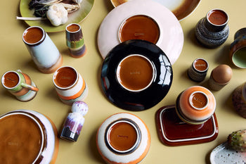 A set of colorful HKLIVING 70s Ceramics Lungo Mugs and other ceramic ware are displayed upside down on a light yellow surface, showcasing their bases with the HKLIVING brand name visible on many pieces.