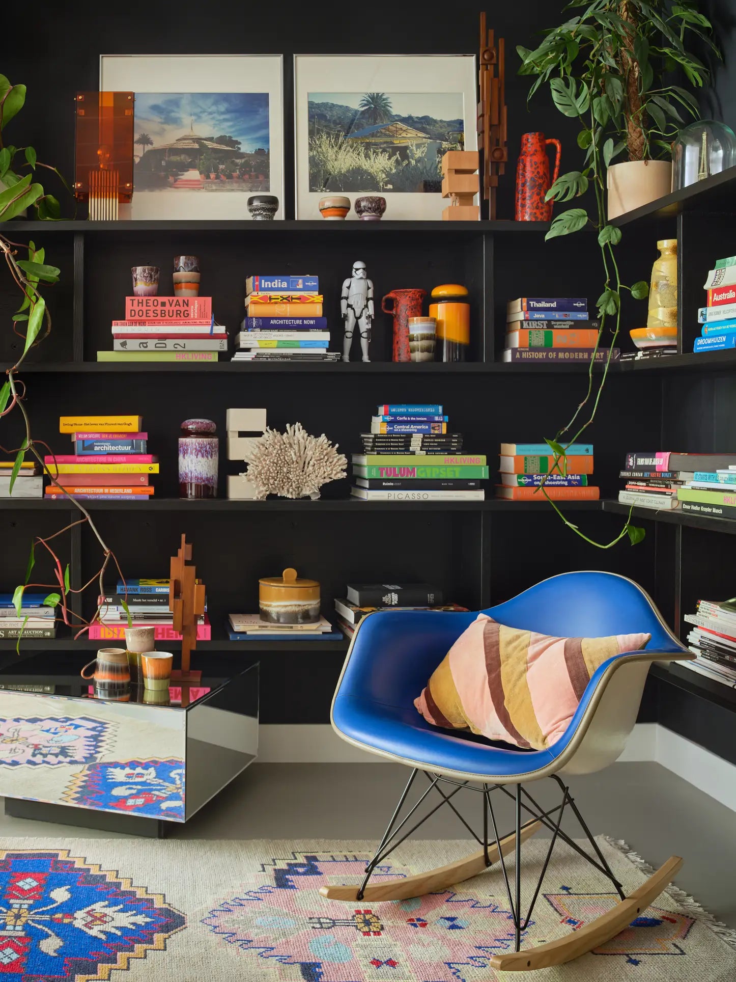 A modern room with a blue rocking chair, striped pillow, colorful rug, and black shelves displaying books, plants, framed art, and HKLIVING 70s Ceramics: Lungo Mugs (Set of 2) creates a cozy, eclectic reading nook with vintage charm.
