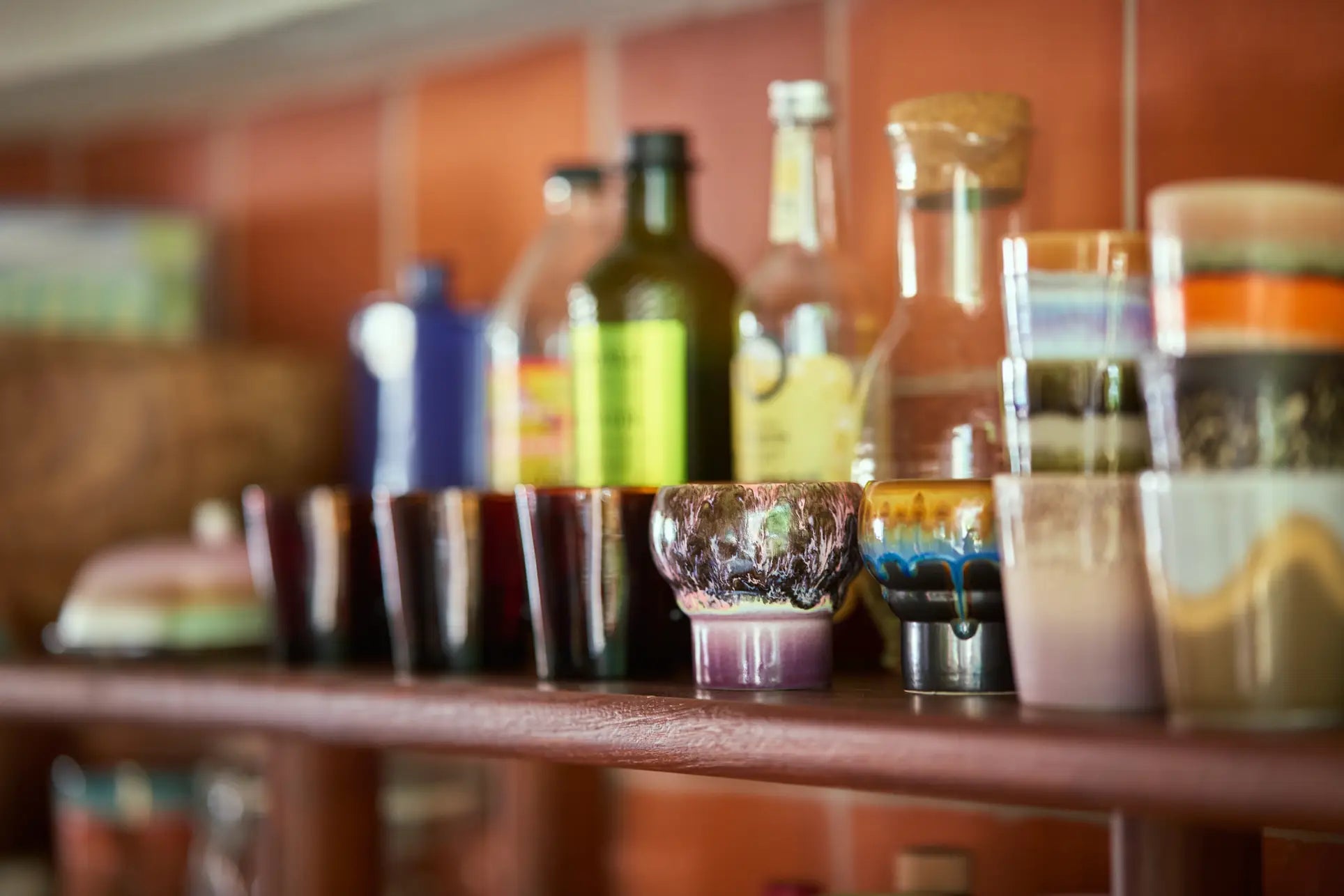 A close-up of HKLIVING 70s Ceramics: Lungo Mugs (Set of 2) on a wooden shelf, with colorful glassware and bottles in the foreground and jars against a reddish tiled wall blurred in the background.