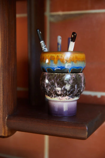 Two HKLIVING 70s Ceramics Lungo Mugs, Set of 2, are stacked on a dark wooden shelf holding paintbrushes. A red-brown tiled wall in the background adds warmth and vintage charm to the scene.