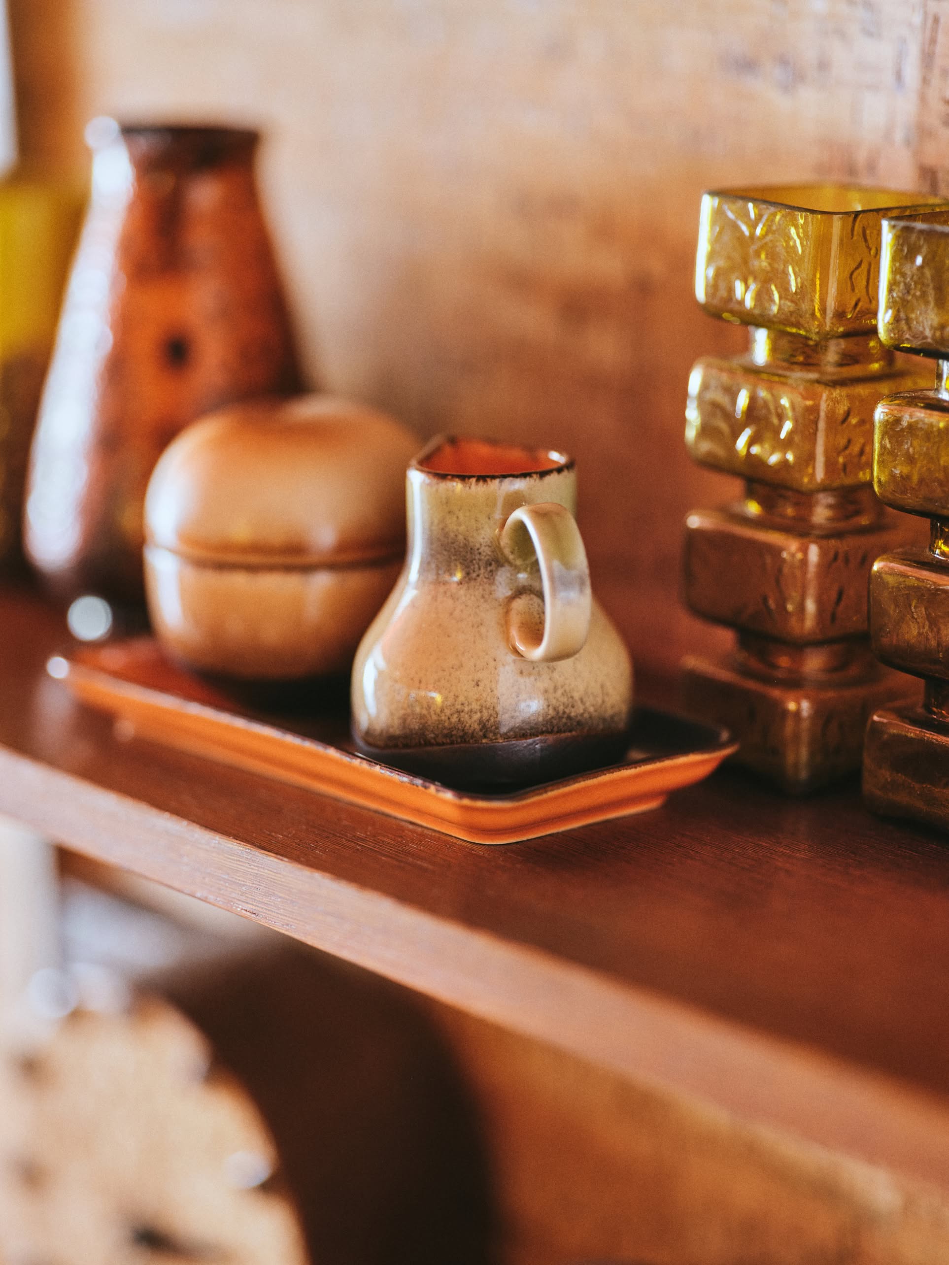 The HKLIVING 70s Ceramics: Milk Jug - 8AM sits on a wooden shelf, accompanied by a ceramic lidded bowl and tray, with shiny gold containers and a textured brown vase nearby.