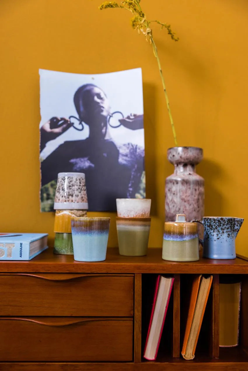 A wooden sideboard displays the HKLIVING 70s Ceramics Milk Jug & Sugar Pot, books, and a tall vase with a dried flower. Behind them, a black-and-white photo is taped to a mustard-yellow wall.