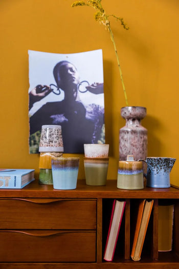 A wooden sideboard displays the HKLIVING 70s Ceramics Milk Jug & Sugar Pot, books, and a tall vase with a dried flower. Behind them, a black-and-white photo is taped to a mustard-yellow wall.