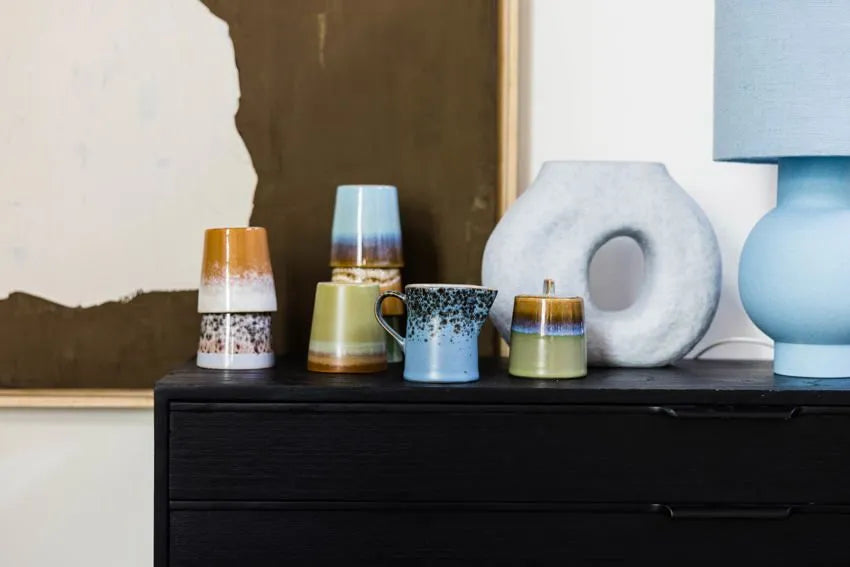 A black dresser displays stacked ceramic mugs in earthy tones, a blue and white mug, the HKLIVING 70s Ceramics: Milk Jug & Sugar Pot, and a modern ring-shaped vase beside a blue lamp, with abstract art in the background.