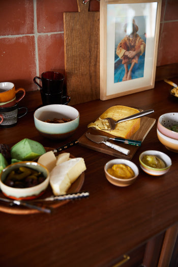 A wooden table showcases cheeses, spreads, and pickled vegetables in HKLIVING 70s Ceramics: Noodle Bowls (Set of 4), with cups, cutlery, and a framed photo against a tiled wall in the background.