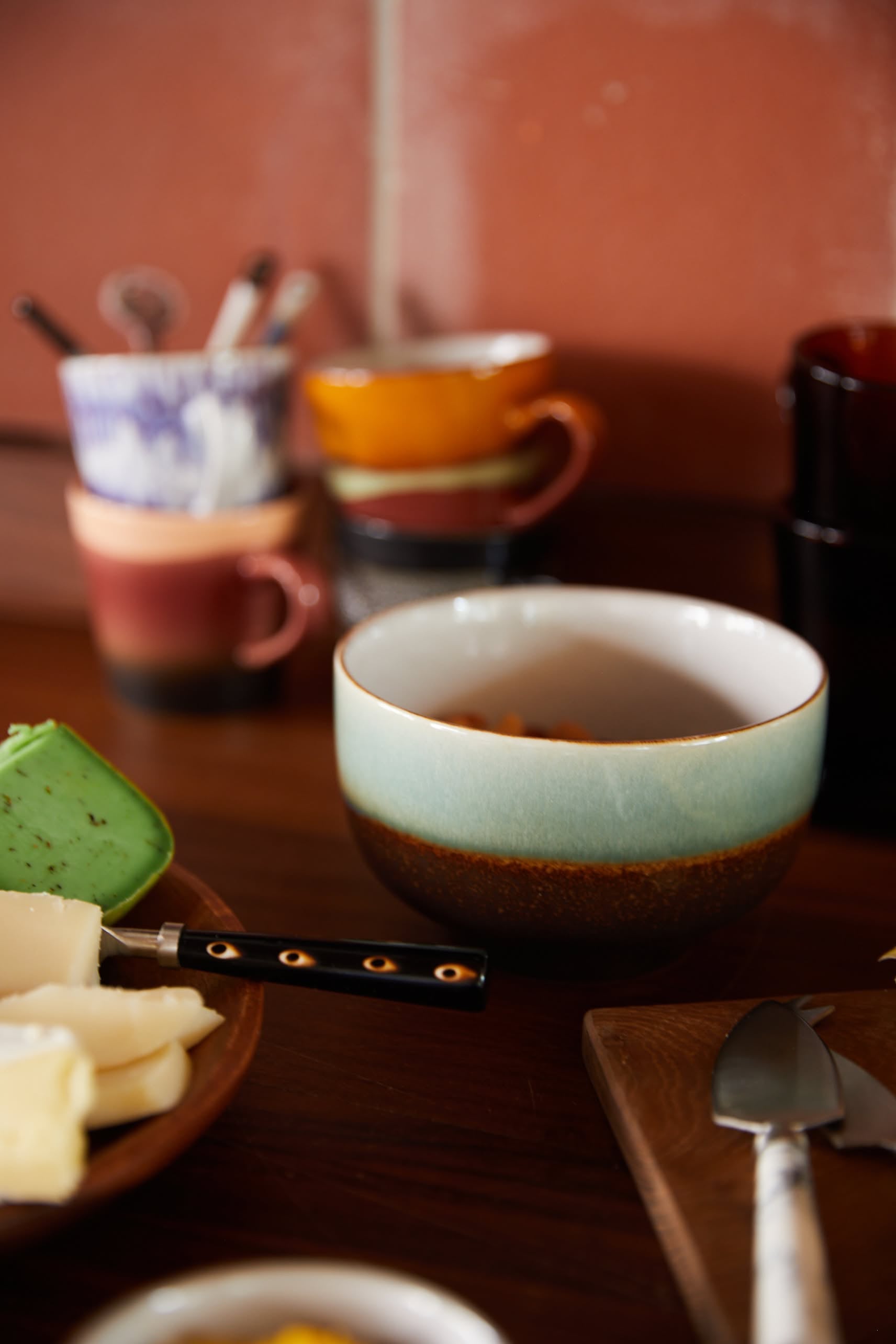 On a wooden table, HKLIVING 70s Ceramics Noodle Bowls (Set of 4) are displayed alongside cheese slices and a black-handled knife, with blurred colorful mugs and utensils in the background evoking vintage retro charm.