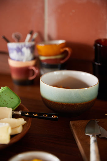 On a wooden table, HKLIVING 70s Ceramics Noodle Bowls (Set of 4) are displayed alongside cheese slices and a black-handled knife, with blurred colorful mugs and utensils in the background evoking vintage retro charm.