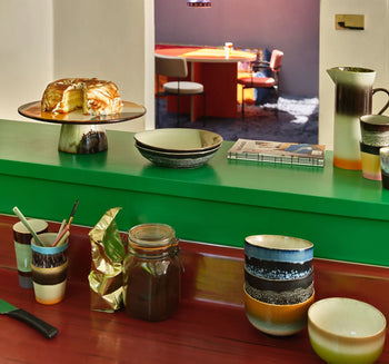 HKLIVING 70s Ceramics: Noodle Bowls, Set of 4, are arranged with cups, a pitcher, and a cake stand on a green and red countertop. A jar, utensils, and book are nearby. In the background, a dining table with chairs is visible through the doorway.