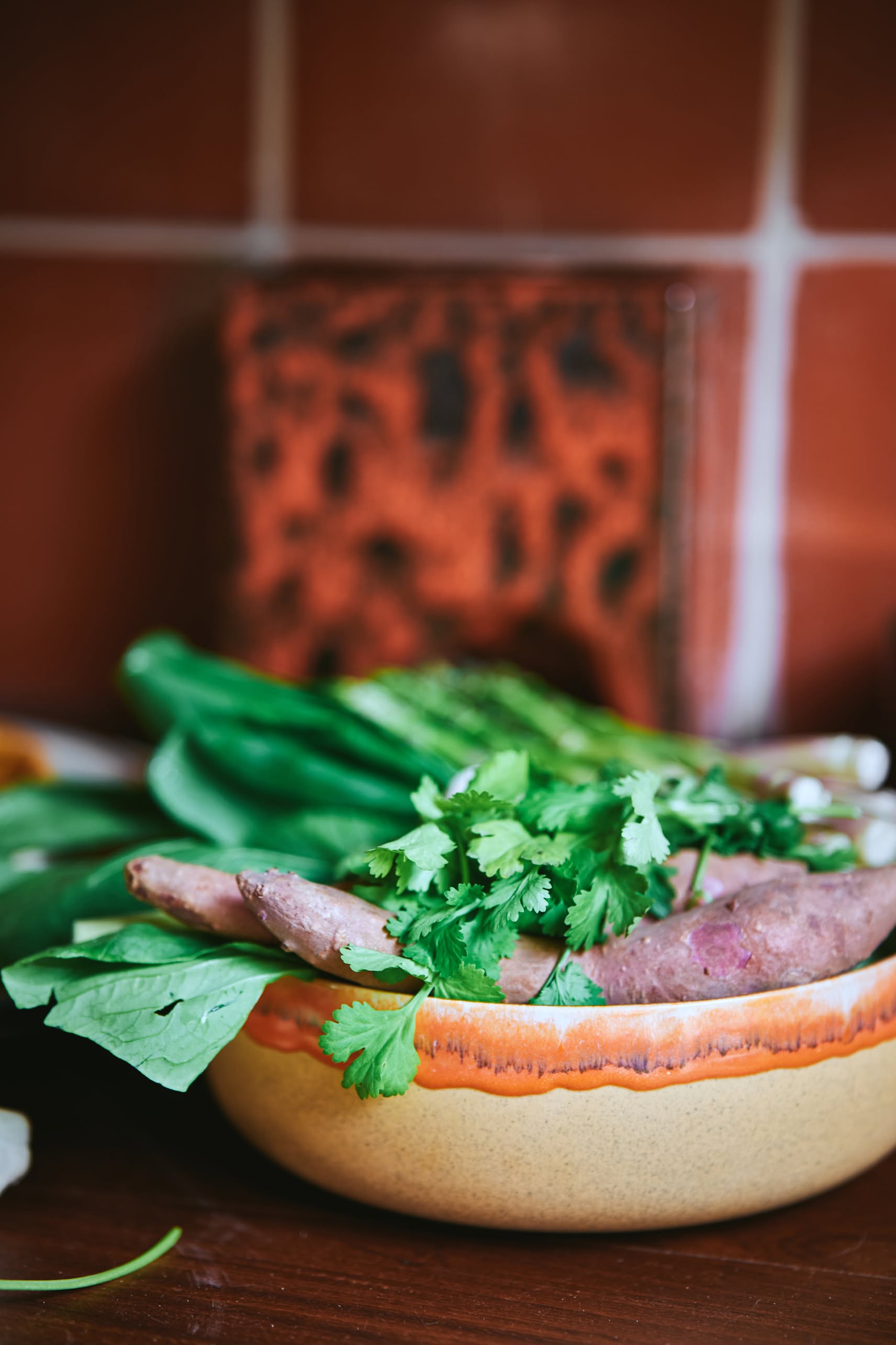 The HKLIVING 70s Ceramics: Salad Bowl by HKLIVING, filled with fresh green vegetables, is placed on a wooden surface in front of a reddish-brown tiled kitchen wall.