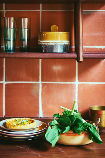 On the counter sits the HKLIVING 70s Ceramics Salad Bowl by HKLIVING, filled with fresh greens. Nearby are stacked plates with a savory pastry, glasses, and a lidded jar. The shelf and reddish-brown tiled walls complete the scene.