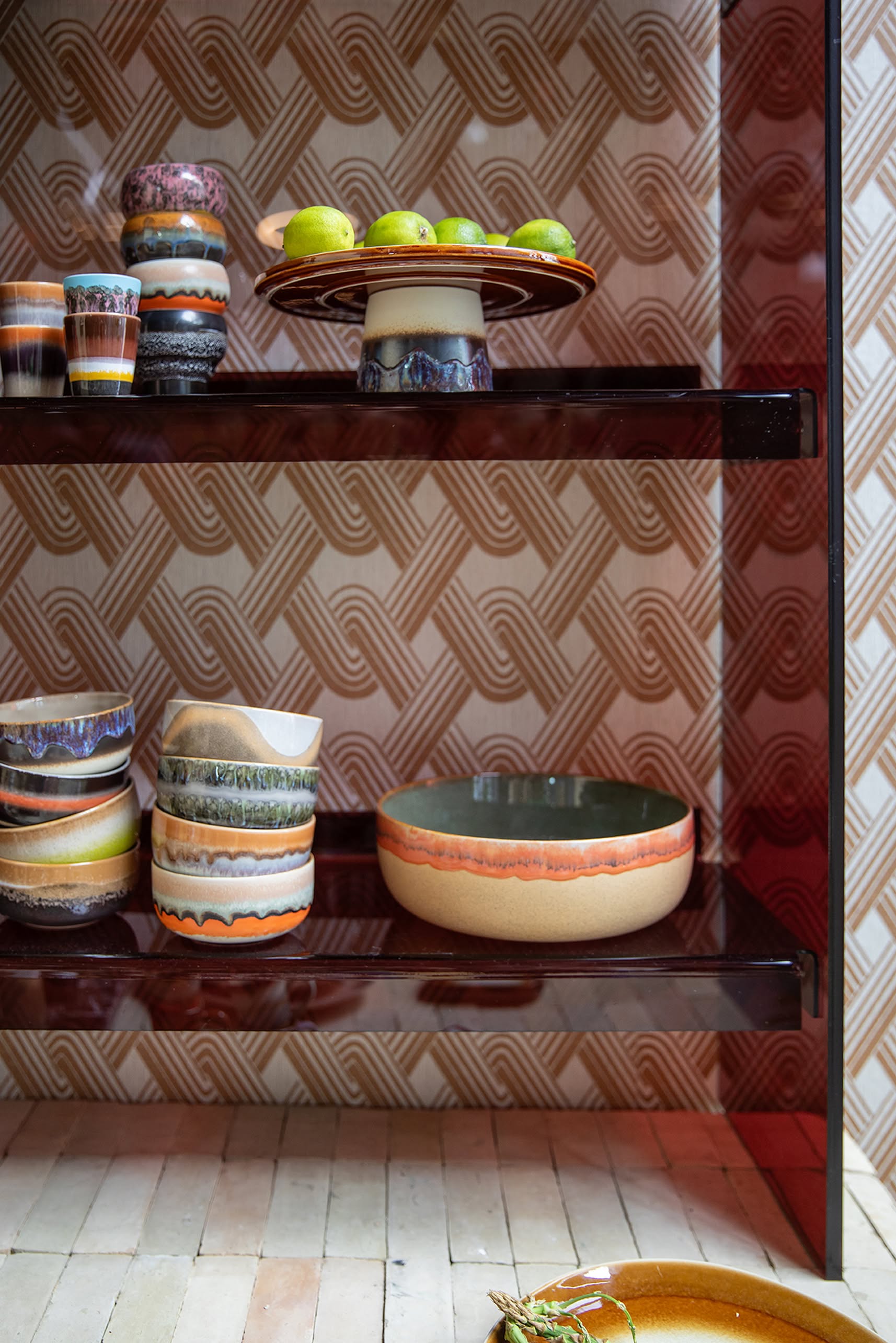 HKLIVING 70s Ceramics Salad Bowls by HKLIVING, with retro earthy patterns, are displayed on shelves—some holding green apples—set against a geometric beige and orange retro-style wallpaper.