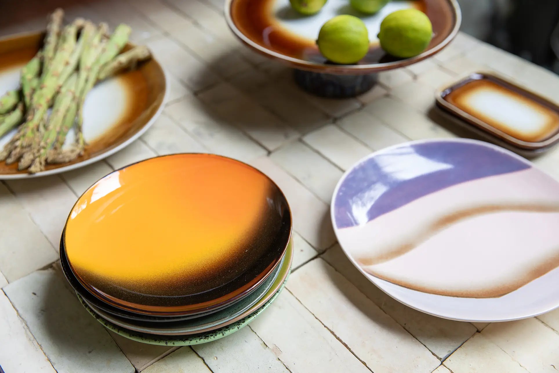 A table set with HKLIVING 70s Ceramics Side Plates (Set of 2), featuring gradient and abstract designs, holds asparagus on one plate and a pedestal dish of green limes. The surface is tiled in a light shade.