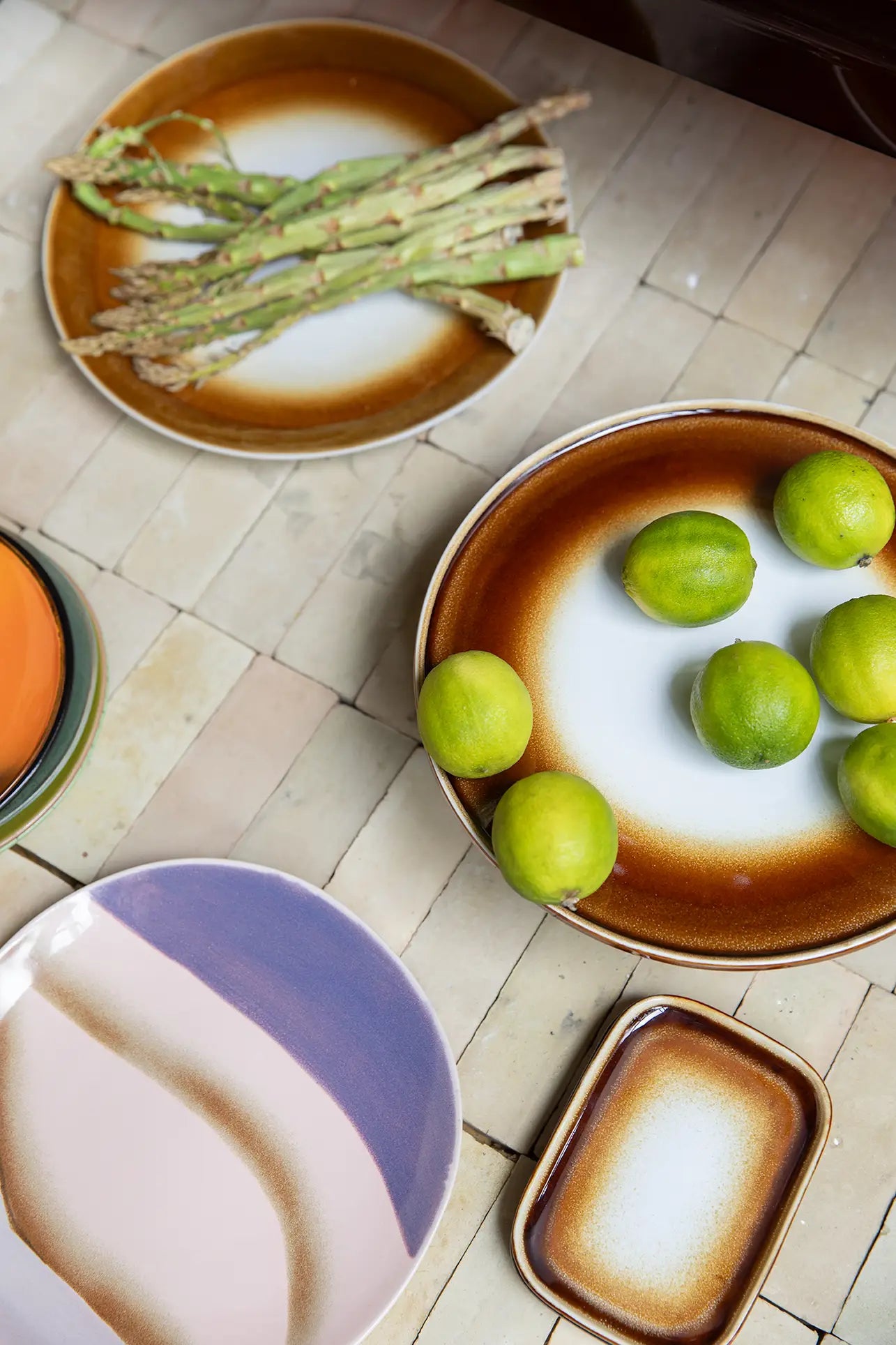 Two colorful HKLIVING 70s Ceramics side plates on a tiled surface—one holds asparagus spears, another fresh limes; both feature abstract retro designs. Set of 2 by HKLIVING.