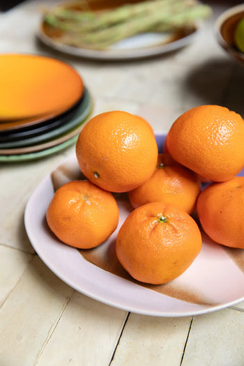 A white plate displays six whole oranges on a light table, with a stack of HKLIVING 70s Ceramics: Side Plates (Set of 2) in the background and blurred asparagus visible behind.