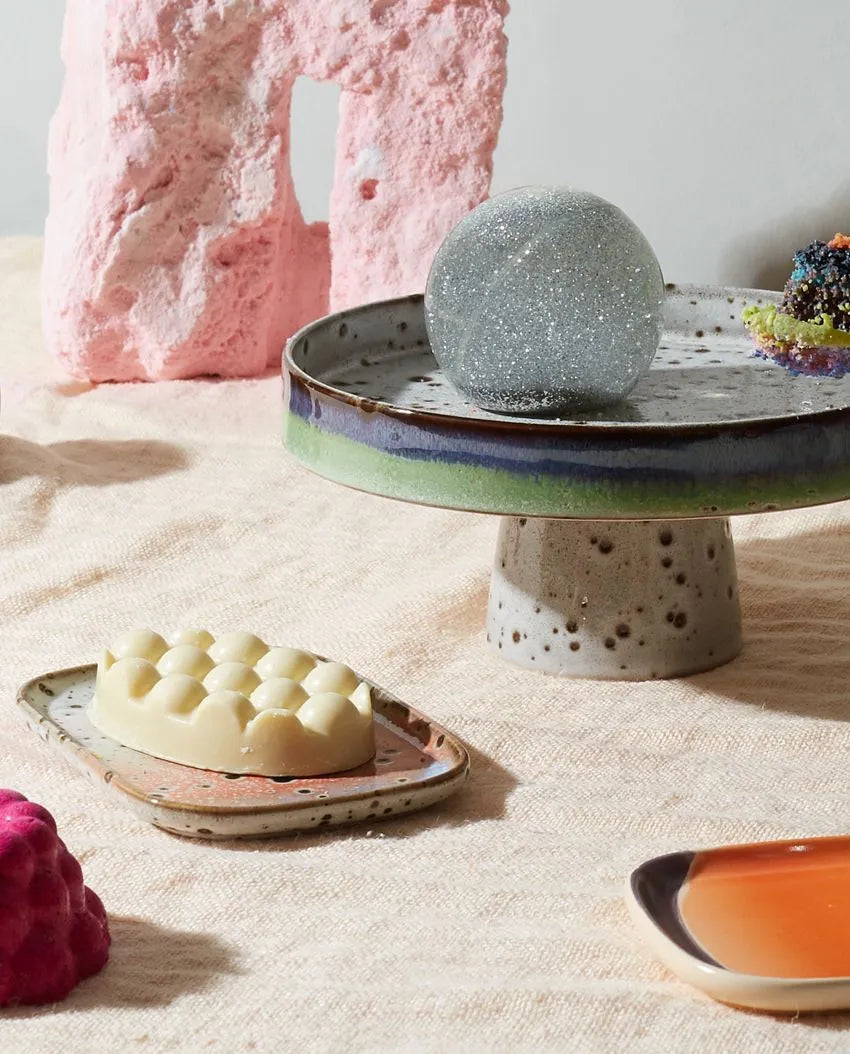 A sparkling, translucent sphere rests on a ceramic cake stand beside colorful desserts and HKLIVING 70s Ceramics: Small Trays, Set of 2 - Atlas by HKLIVING, displayed atop a textured cream tablecloth with a pink arch-shaped decoration in the background.