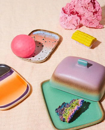 Colorful objects on a beige surface, featuring HKLIVING 70s Ceramics: Small Trays, Set of 2 - Atlas, pastel dishes, a pink sphere, yellow brush, and a slice of multicolored cake on a green plate with lid.