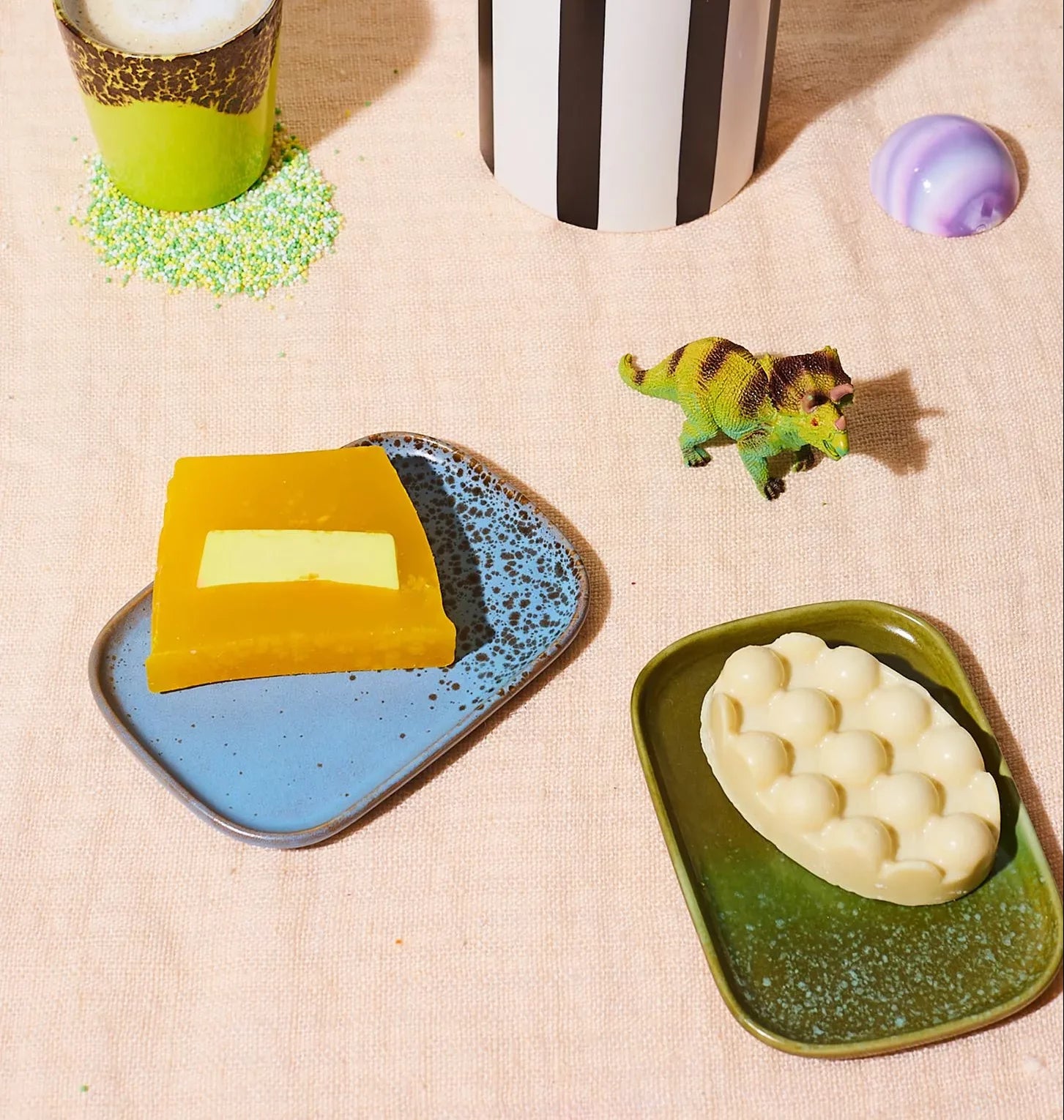 A flat lay of two HKLIVING 70s Ceramics: Small Trays, Set of 2 - Atlas by HKLIVING, with soap bars, a toy dinosaur, a green sprinkle cup, a striped cup, and a round purple object on light beige fabric.