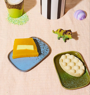 A flat lay of two HKLIVING 70s Ceramics: Small Trays, Set of 2 - Atlas by HKLIVING, with soap bars, a toy dinosaur, a green sprinkle cup, a striped cup, and a round purple object on light beige fabric.