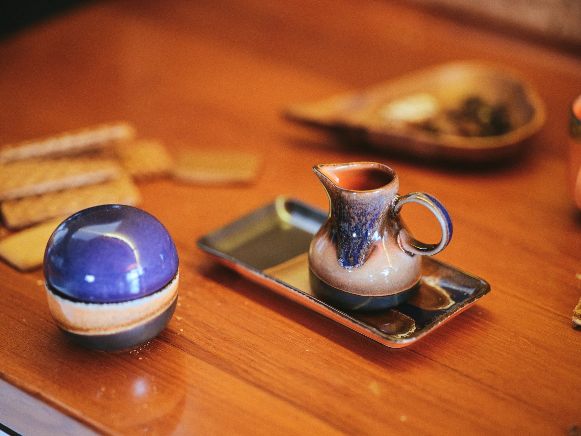 On a wooden table, the HKLIVING 70s Ceramics: Small Trays, Set of 2 - Twenty Four Hours display a small ceramic pitcher, a round lidded blue container with retro 70s charm, and some biscuits.