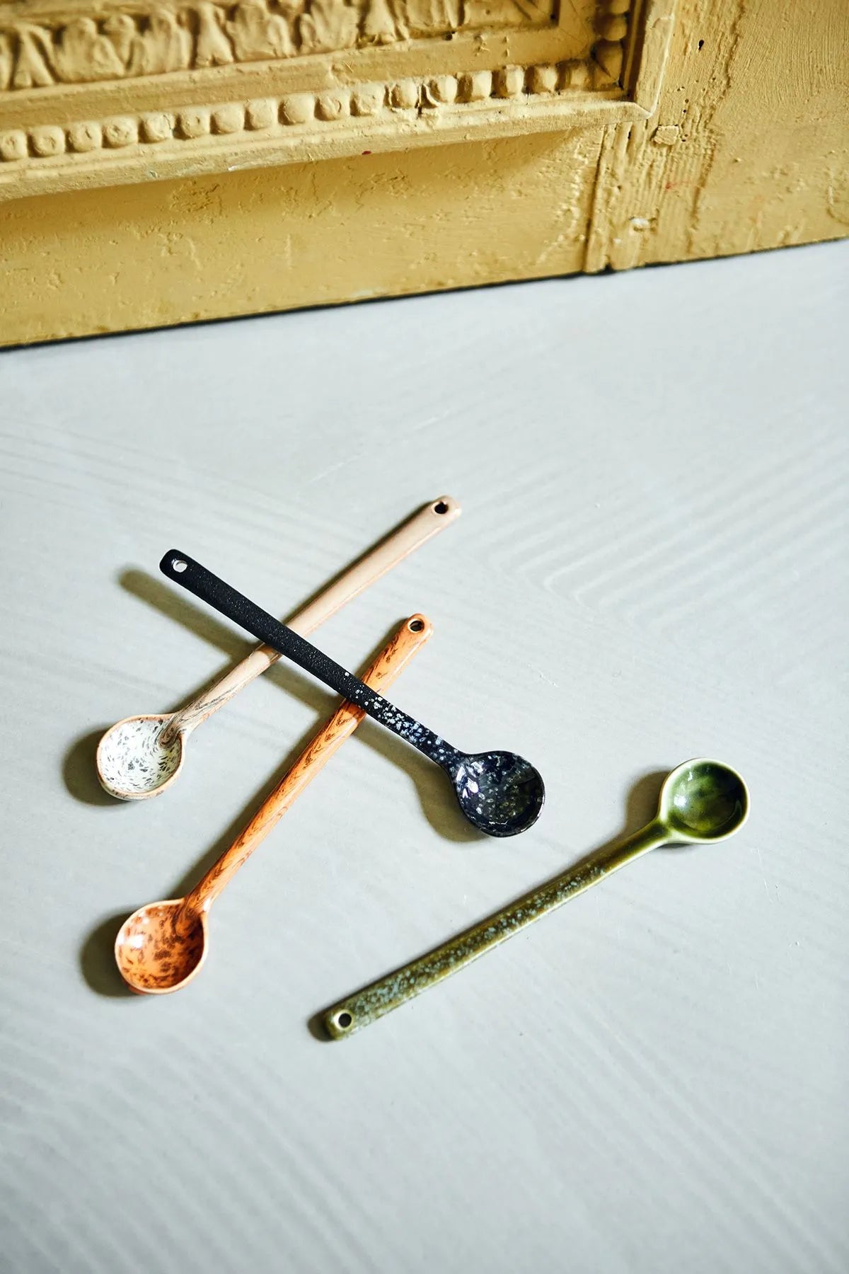 The HKLIVING 70s Ceramics: Spoons L, Set of 4 features black, gold, copper, and silver spoons arranged on a textured surface near a decorative gold frame, capturing the retro charm of the 70s.