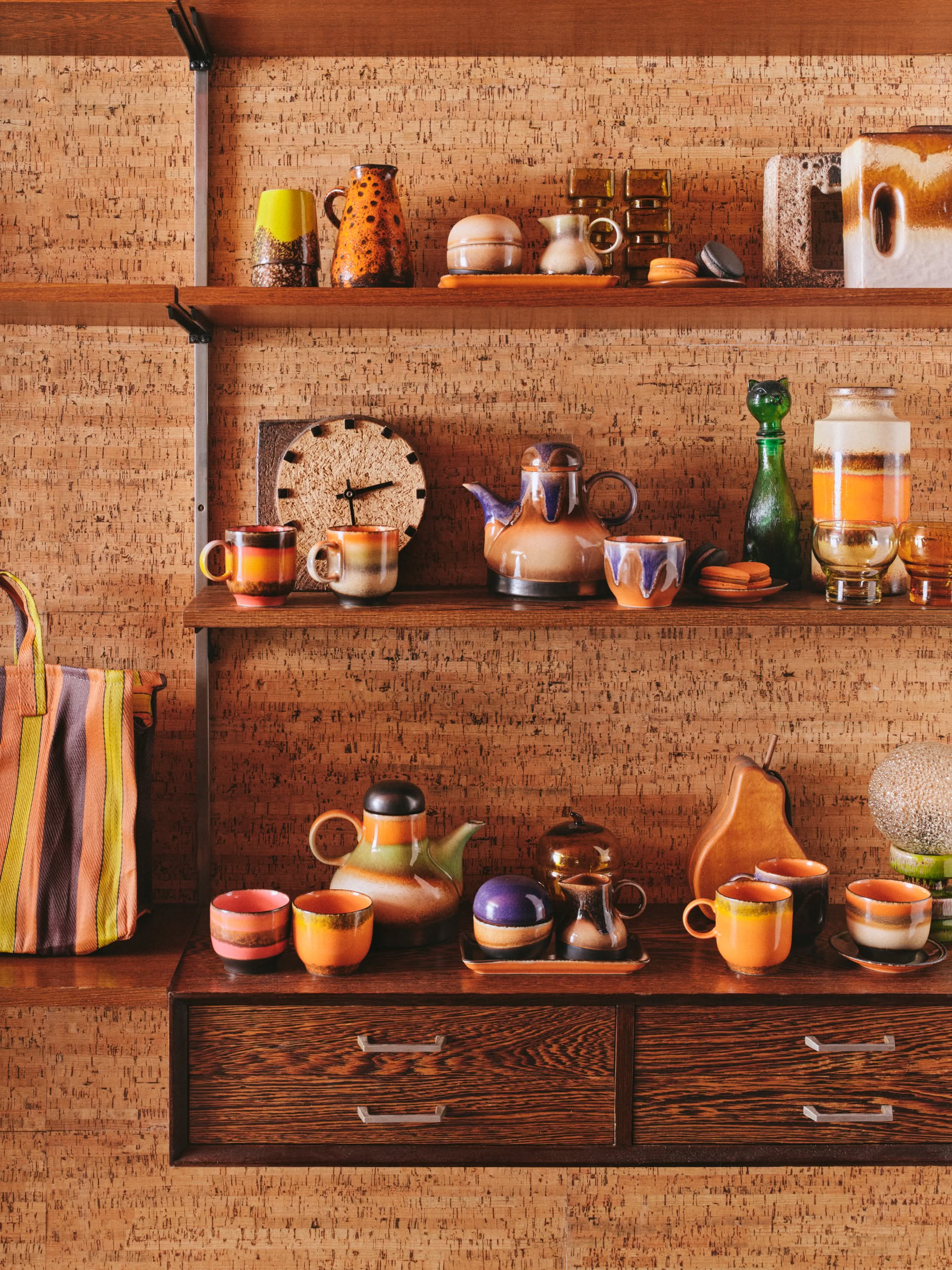 Wooden shelves display colorful ceramic teapots, mugs, the HKliving 70s Ceramics: Sugar Pot for condiments, jars, bottles, and a striped tote bag against a textured tan wall for an eclectic retro vibe.