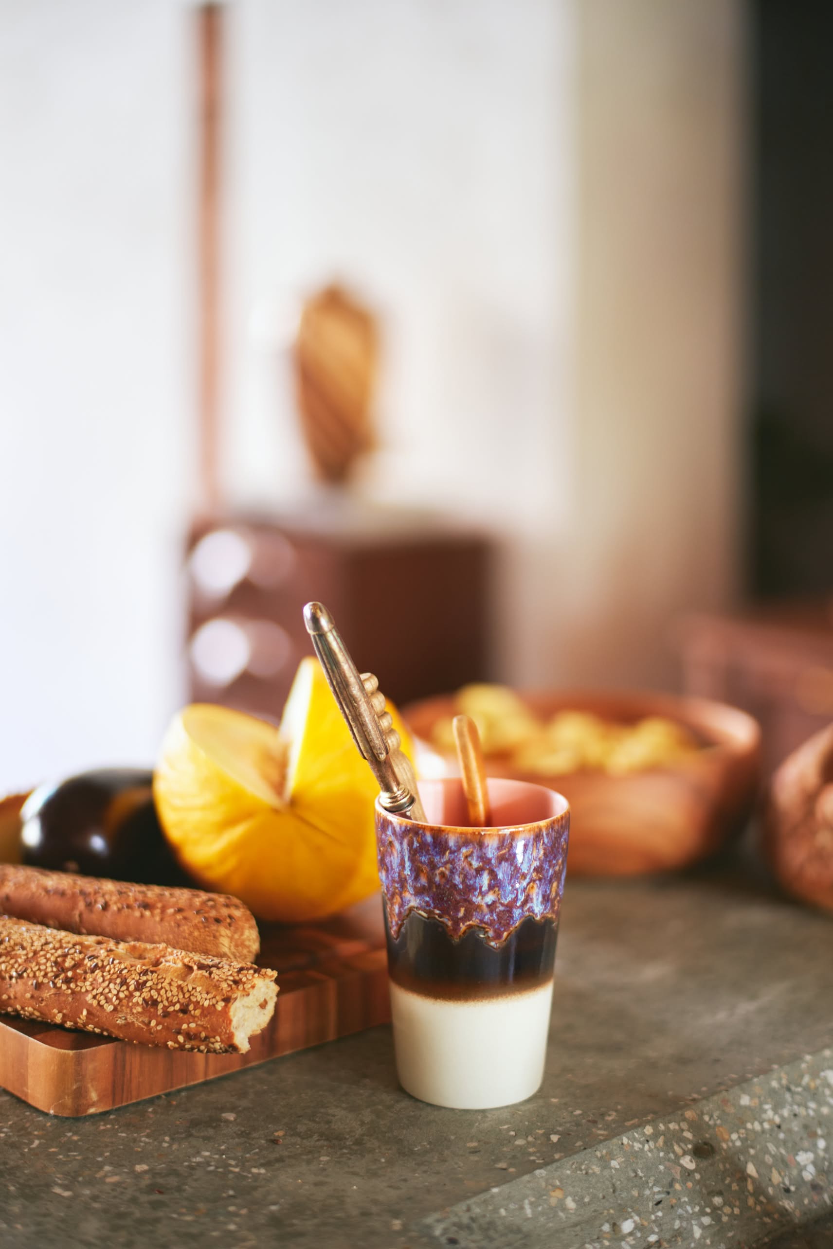 The HKLIVING 70s Ceramics Tea Mugs, Set of 2, sit on a counter with seeded breadsticks, sliced melon, and a fruit bowl in a warmly lit kitchen.