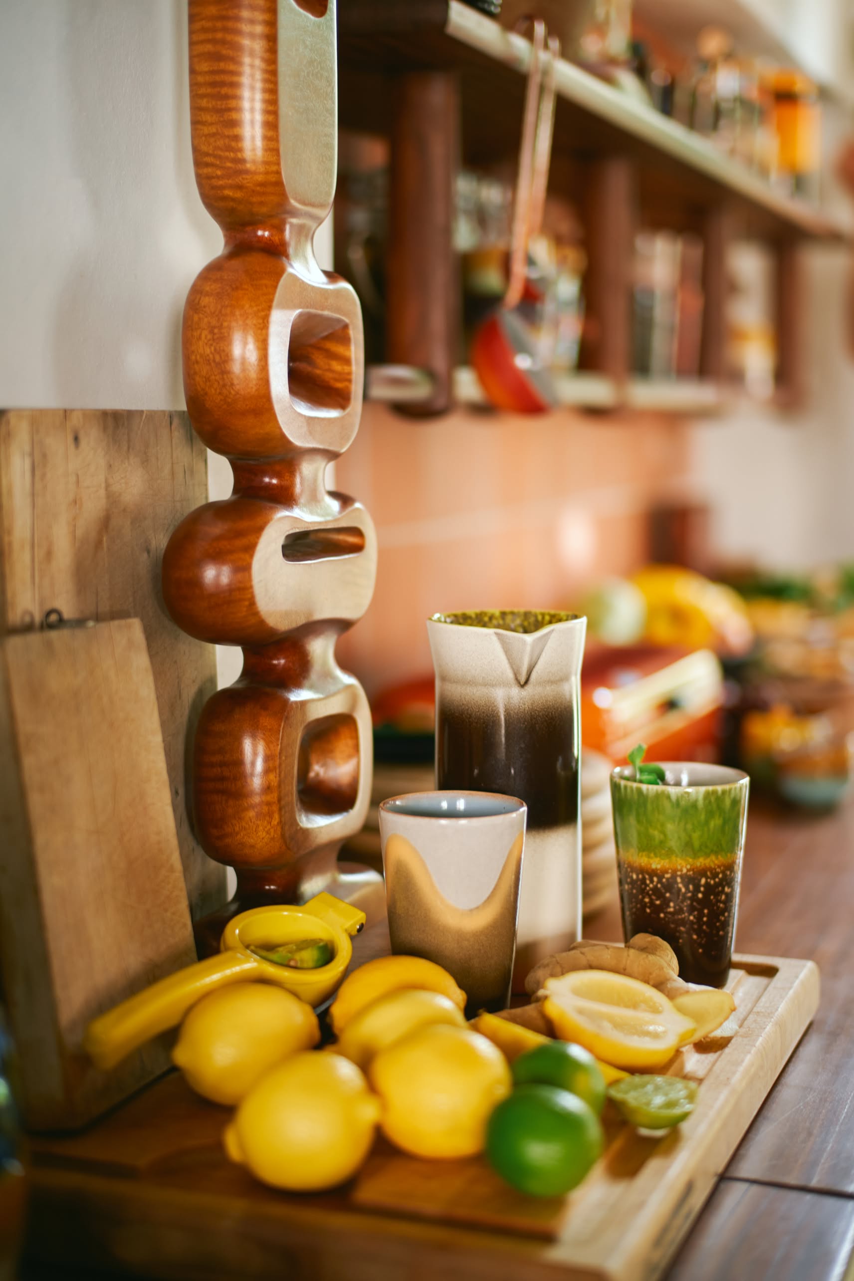 On the kitchen counter, a wooden cutting board with lemons, a lime, a banana, and a glass of chia drink sits beside the HKLIVING 70s Ceramics: Tea Mugs (set of 2) and a modern wooden sculpture; kitchen shelves appear in the background.