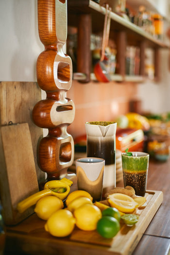On the kitchen counter, a wooden cutting board with lemons, a lime, a banana, and a glass of chia drink sits beside the HKLIVING 70s Ceramics: Tea Mugs (set of 2) and a modern wooden sculpture; kitchen shelves appear in the background.