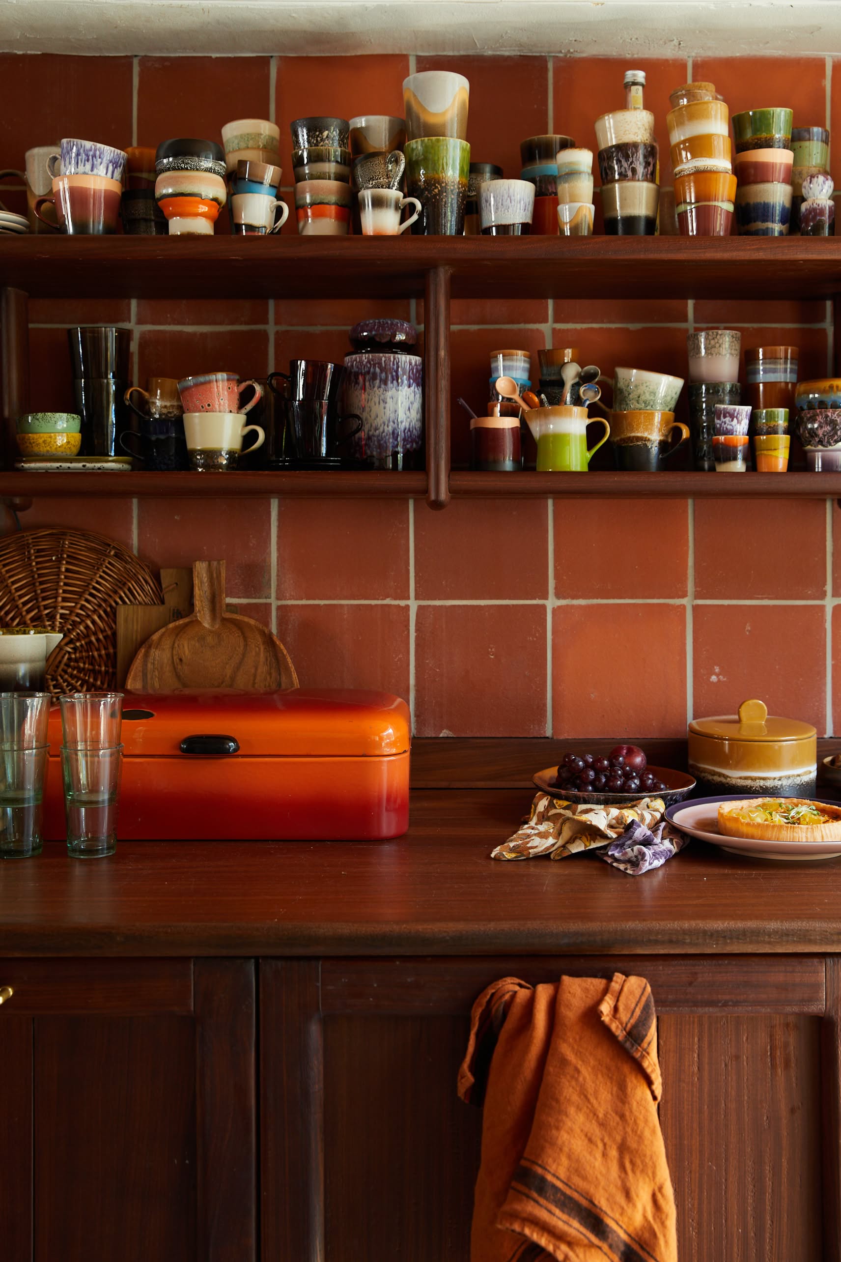 A cozy kitchen with reddish-brown tile walls, wooden shelves displaying HKLIVING 70s Ceramics Tea Mugs (Set of 2), a wooden counter with an orange bread box, glassware, dishes of food and fruit, and an orange towel hanging from a drawer.