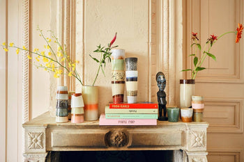 A charming vintage vibe is created by HKliving 70s Ceramics Tea Mugs, vases of red and yellow flowers, stacked books, and a black statue atop a mantel against an ornately carved cream wall.